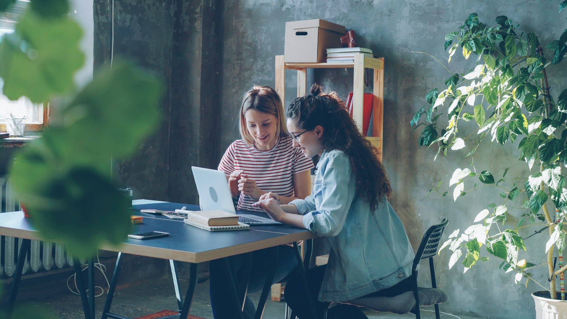 Two women collaborate at a desk with a laptop.